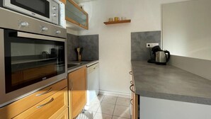 A compact kitchen with a built-in oven, microwave, and dishwasher. There is a wooden cabinet with drawers and a countertop with a kettle.