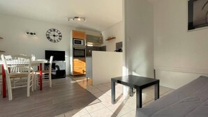 A modern living room with a grey sofa, a dining table with chairs, a kitchen area with appliances, and a wall clock.