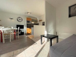 A modern living room with a grey sofa, a dining table with chairs, a kitchen area with appliances, and a wall clock. - 200m from resort center, 1st floor, balcony, tv, 29m², Serre Chevalier (Briançon)