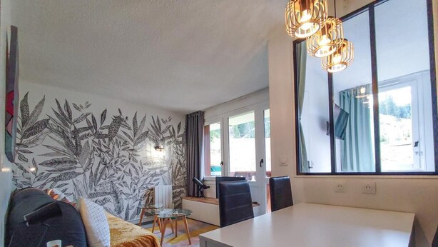 A modern room with a bed, a dining table, chairs, and a large window.