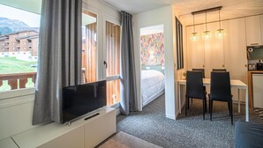A hotel room with a TV, a bed, a dining area with chairs, a small table, and a view of a building and a grassy area.