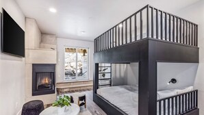 A modern bedroom with a bunk bed, a fireplace, a TV, and a window with a view of trees.