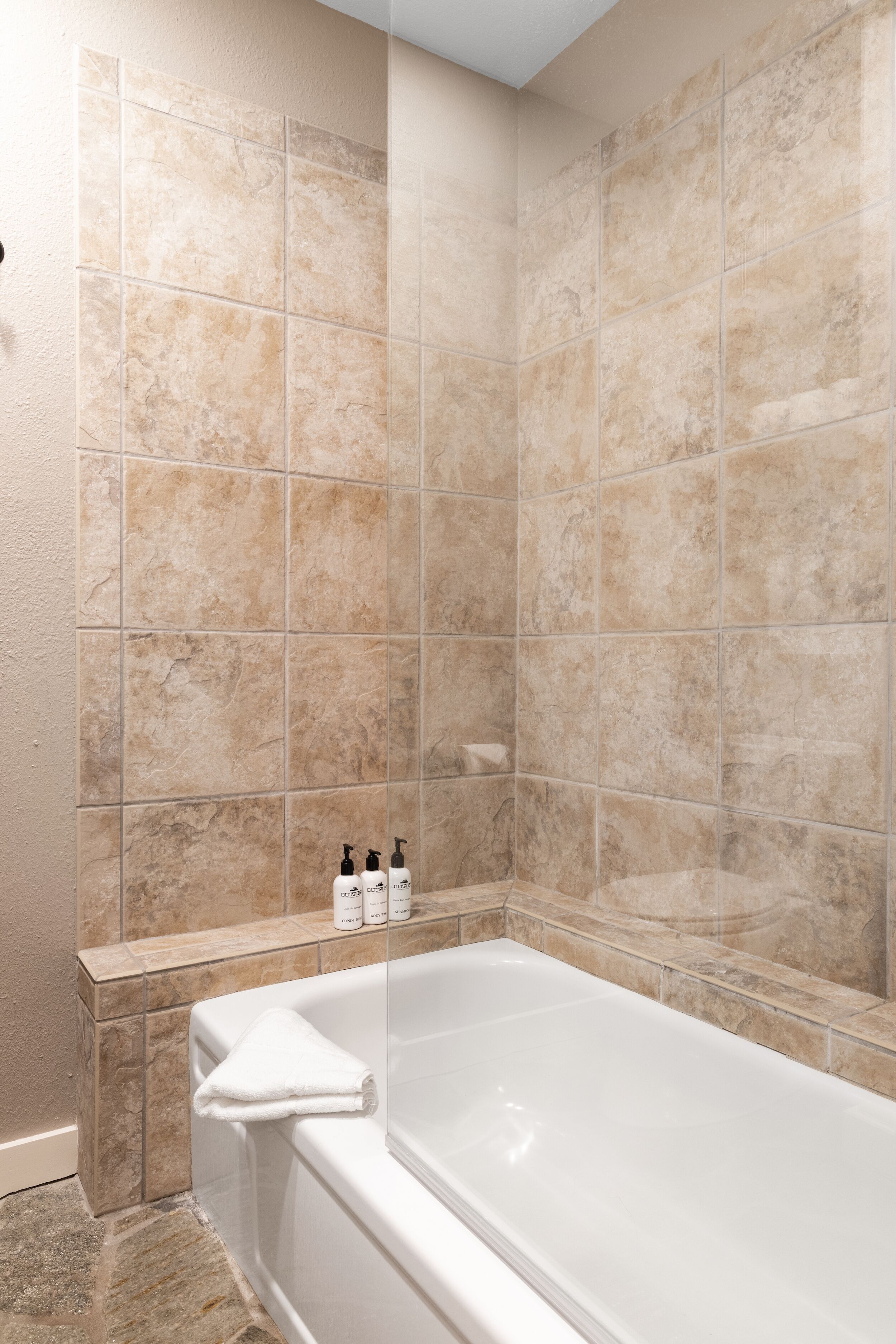 A bathroom with beige tiles, a bathtub, and a shelf with toiletries.
