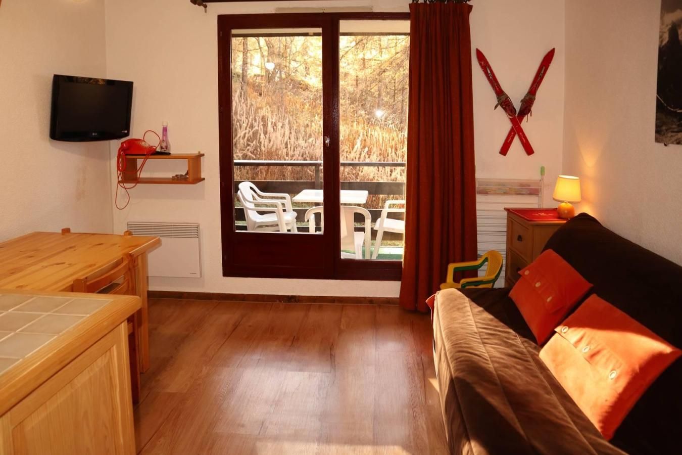 A room with a wooden table, a sofa with red cushions, a television mounted on the wall, and a sliding glass door leading to an outdoor seating area.
