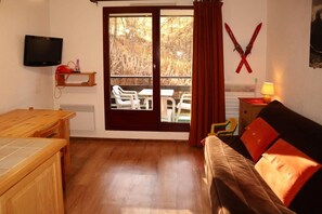 A room with a wooden table, a sofa with red cushions, a television mounted on the wall, and a sliding glass door leading to an outdoor seating area.