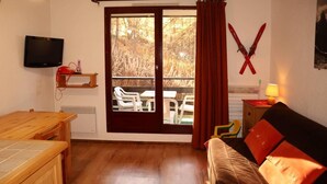 A room with a wooden table, a sofa with red cushions, a TV mounted on the wall, and a sliding glass door leading to an outdoor seating area.