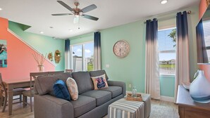 A living room with a gray sofa, striped ottoman, ceiling fan, and a large window with curtains. - Surfstyle At Margaritaville By Shine Villas 022 3 Bedroom Villa (Kissimmee)