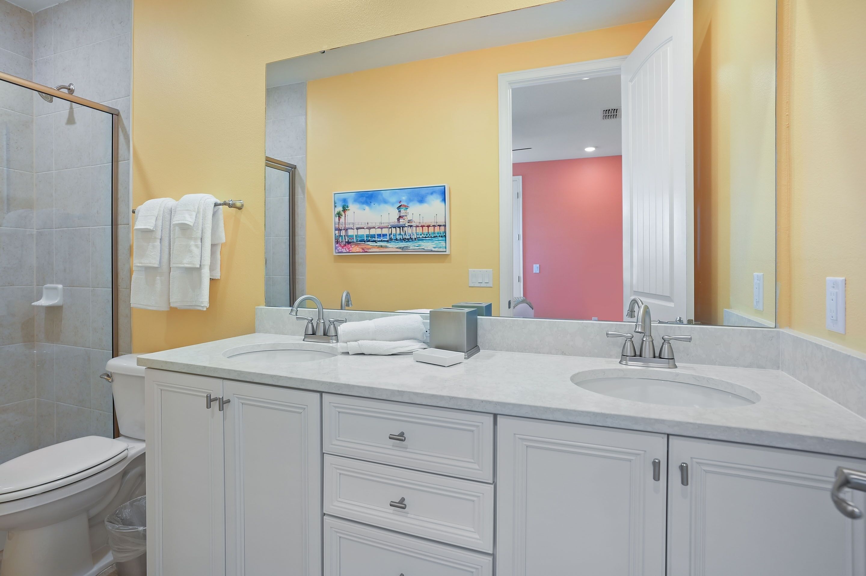 A bathroom with a double sink, white cabinets, a glass shower enclosure, and a painting on the wall.