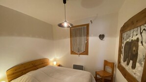 A bedroom with a wooden bed, bedside lamps, a chair, a window with a curtain, and a bear painting on the wall. - 150m from ski slopes, 5th floor, view resort, balcony, tv, 55m², Méribel (Méribel - Les Allues)