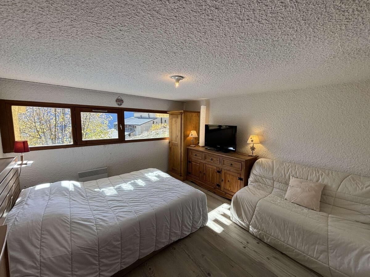 Ein Schlafzimmer mit Bett, einer Couch, einem Fernseher auf einer Holzkommode und einem Fenster mit Blick auf Bäume.