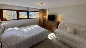 Ein Schlafzimmer mit Bett, einer Couch, einem Fernseher auf einer Holzkommode und einem Fenster mit Blick auf Bäume.