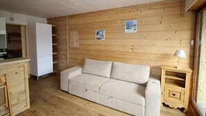 Ein gemütliches Zimmer mit Holzwand, einem beigefarbenen Sofa, einem kleinen Nachttisch mit Lampe und einem weißen Kühlschrank.