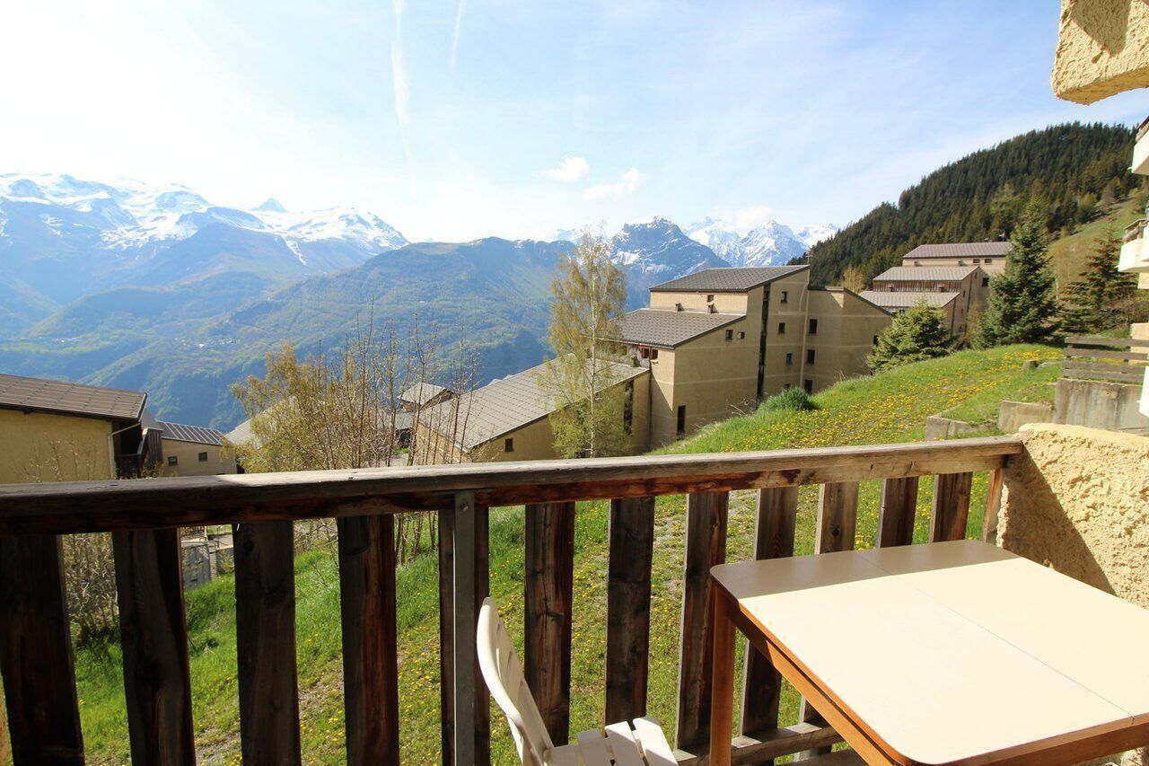 Eine Holzdielen-Terrasse mit Tisch und Stuhl, mit Blick auf ein Dorf, das von Gebäuden und schneebedeckten Bergen gesäumt wird.