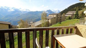 Eine Holzdielen-Terrasse mit Tisch und Stuhl, mit Blick auf ein Dorf, das von Gebäuden und schneebedeckten Bergen gesäumt wird.