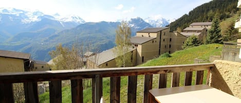 Una terraza de madera con una mesa y una silla, con vistas a un pueblo con edificios y montañas nevadas al fondo.
