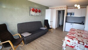 A modern living room with a grey sofa, two armchairs, a dining table with a red and white tablecloth, a kitchen area, and a painting on the wall. - Ski-in/ski-out & foot of the slopes, 3rd floor, view mountain, balcony, tv, ski locker, 55m² (Auris)