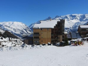 A multi-story building with a wooden facade and a snow-covered roof, surrounded by a snowy landscape and mountains in the background. - 10m from ski slopes, 2nd floor, balcony, tv, ski locker, 23m², Auris en Oisans (Auris)