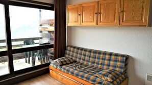 Een kleine kamer met een geruite bank, houten kasten, een groot raam en uitzicht op tuinmeubilair.