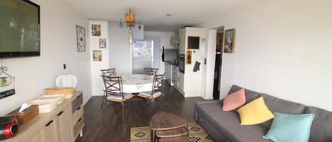A compact living space with a dining area, kitchenette, and a gray sofa with colorful cushions.