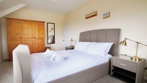Un letto rifatto con lenzuola e asciugamani bianchi, una testiera grigia rivestita in tessuto, un comodino con una lampada e un armadio in legno.