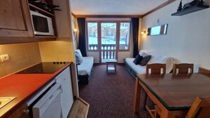 A compact kitchen with a microwave, stove, and dishwasher. There is a dining area with a table and chairs, and a sofa. A balcony with a railing is visible through the window. - 50m from ski slopes, 4th floor, view ski slopes, balcony, parking, tv, 29m², La Plagne (La Plagne)