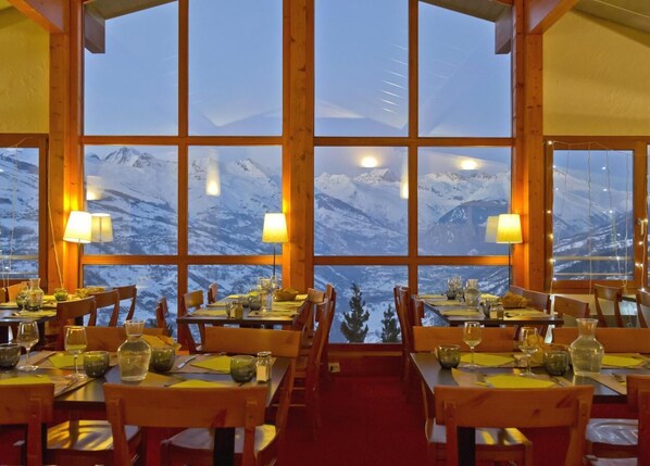 Un restaurant doté de nombreuses tables et chaises, avec de grandes fenêtres offrant une vue sur des montagnes enneigées.