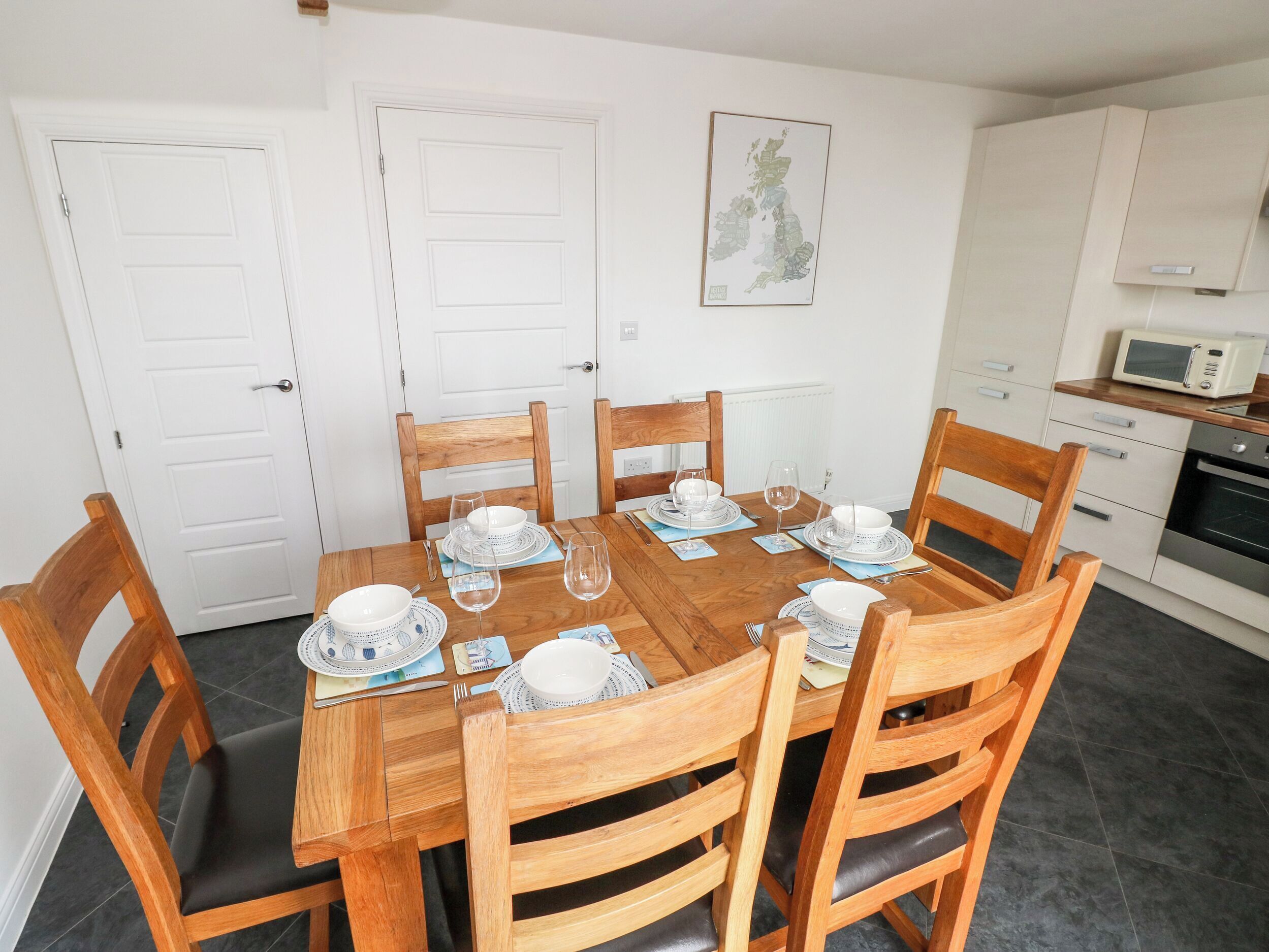A dining area with a wooden table set for six, surrounded by wooden chairs. There are white doors and a kitchen area with a microwave and oven visible.