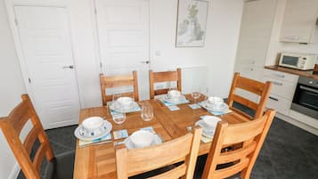 A dining area with a wooden table set for six, surrounded by wooden chairs. There are white doors and a kitchen area with a microwave and oven visible.