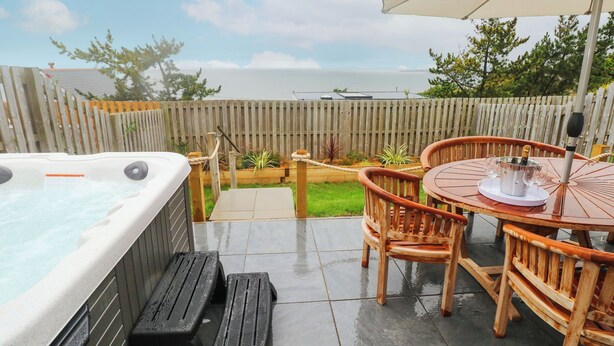 A hot tub, wooden deck, and outdoor seating area with a view of the sea. - Tŷ Glas (Bridgend)