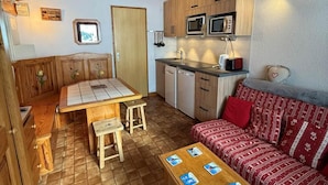 Et kompakt køkken med træskabe, en mikrobølgeovn og en vask. Der er et spiseområde med et bord og stole, samt en sofa med mønstrede puder.