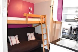 Ein Stockbett mit Holzleiter, ein dunkelbraunes Sofa mit gestreiften Kissen, ein weißes Waschbecken und ein Küchenbereich mit hängendem Geschirr.