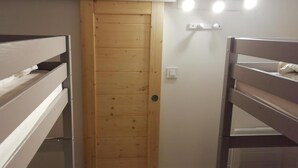 Ein schmaler Raum mit zwei Etagenbetten, einer Holztür und einem Lichtschalter an der Wand.
