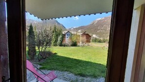A view from a room with a door open, revealing a patio with a pink chair and a grassy area with buildings and mountains in the background. - 100m from resort center, Ground floor, terrace, tv, ski locker, 42m², Serre Chevalier (La Salle-les-Alpes)