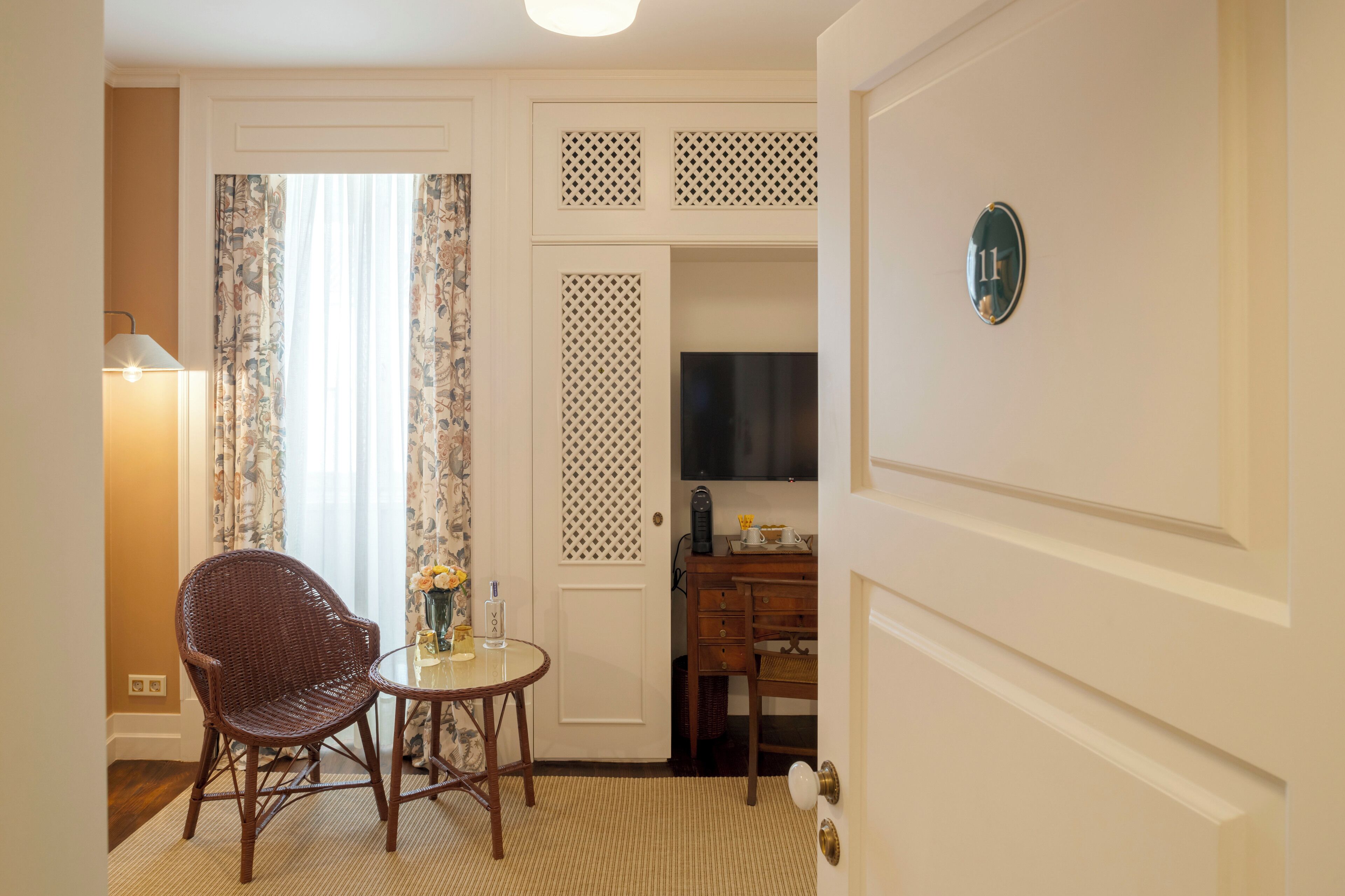 Une chambre d’hôtel avec un fauteuil en osier, une petite table ronde, une lampe, un téléviseur et un bureau avec des tiroirs.