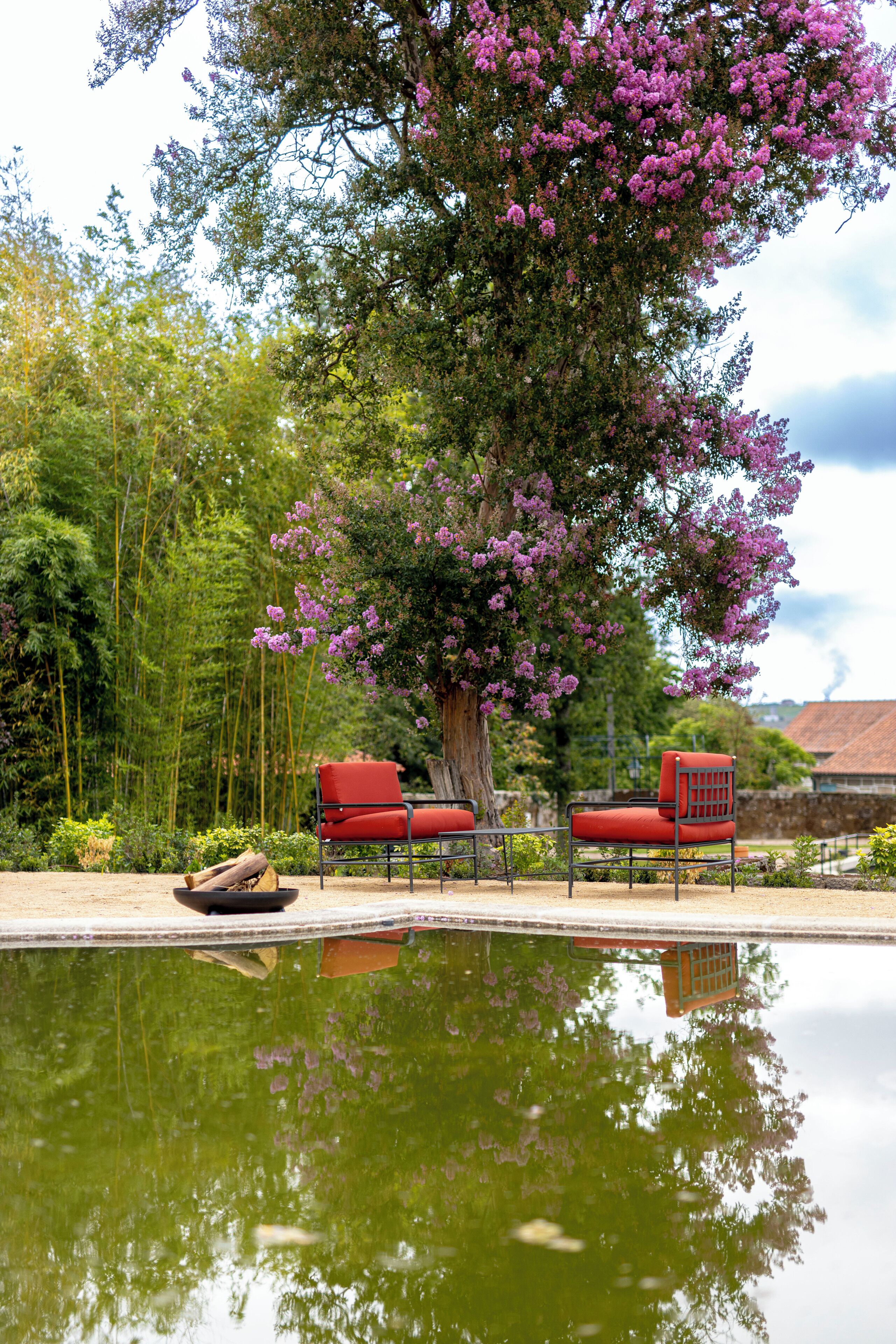 A serene outdoor area with a reflective pool, two red chairs, and a large tree with purple flowers.