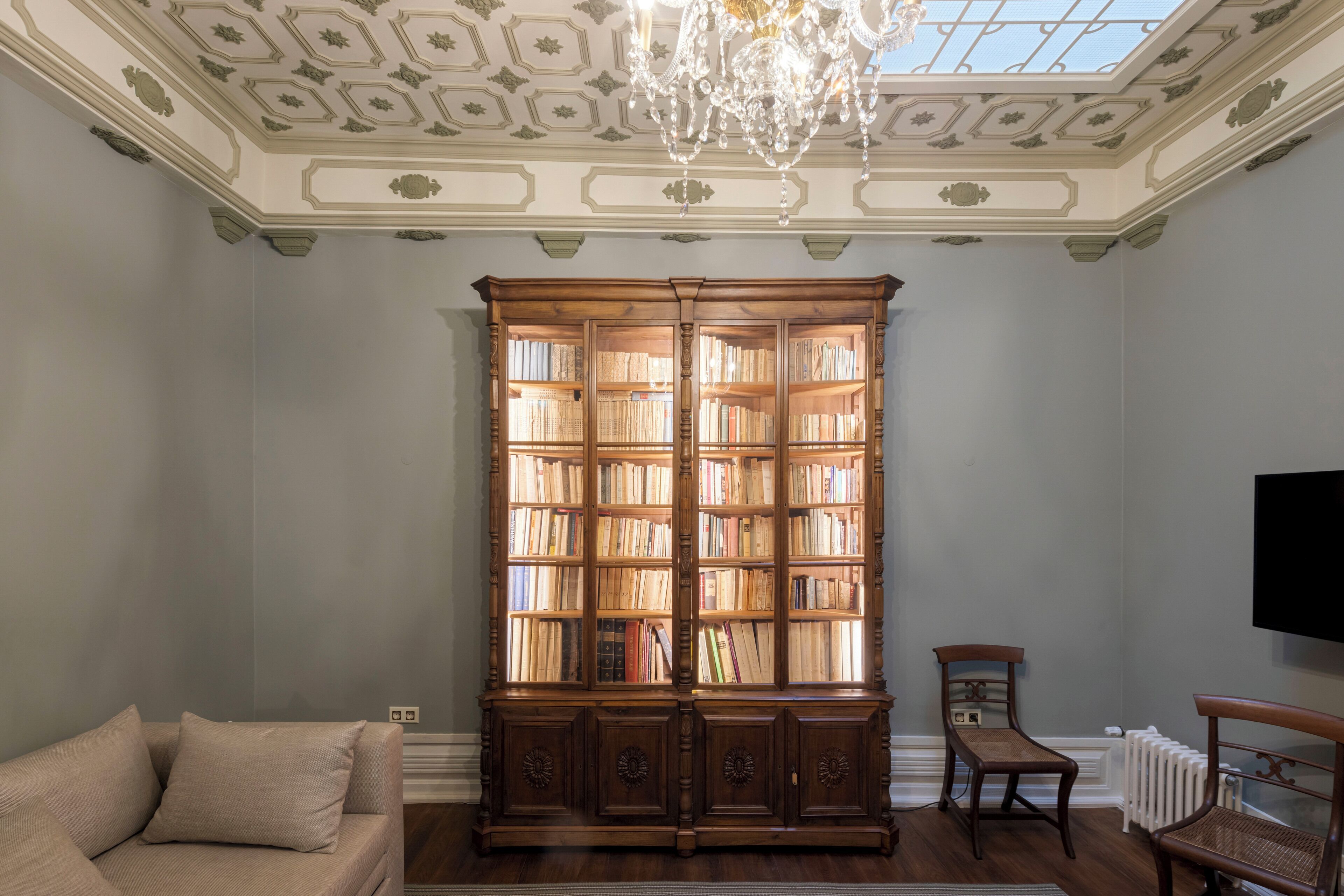 Una habitación con una gran estantería de madera llena de libros, un sofá color beige, dos sillas de madera, una araña y un televisor de pantalla plana montado en la pared.