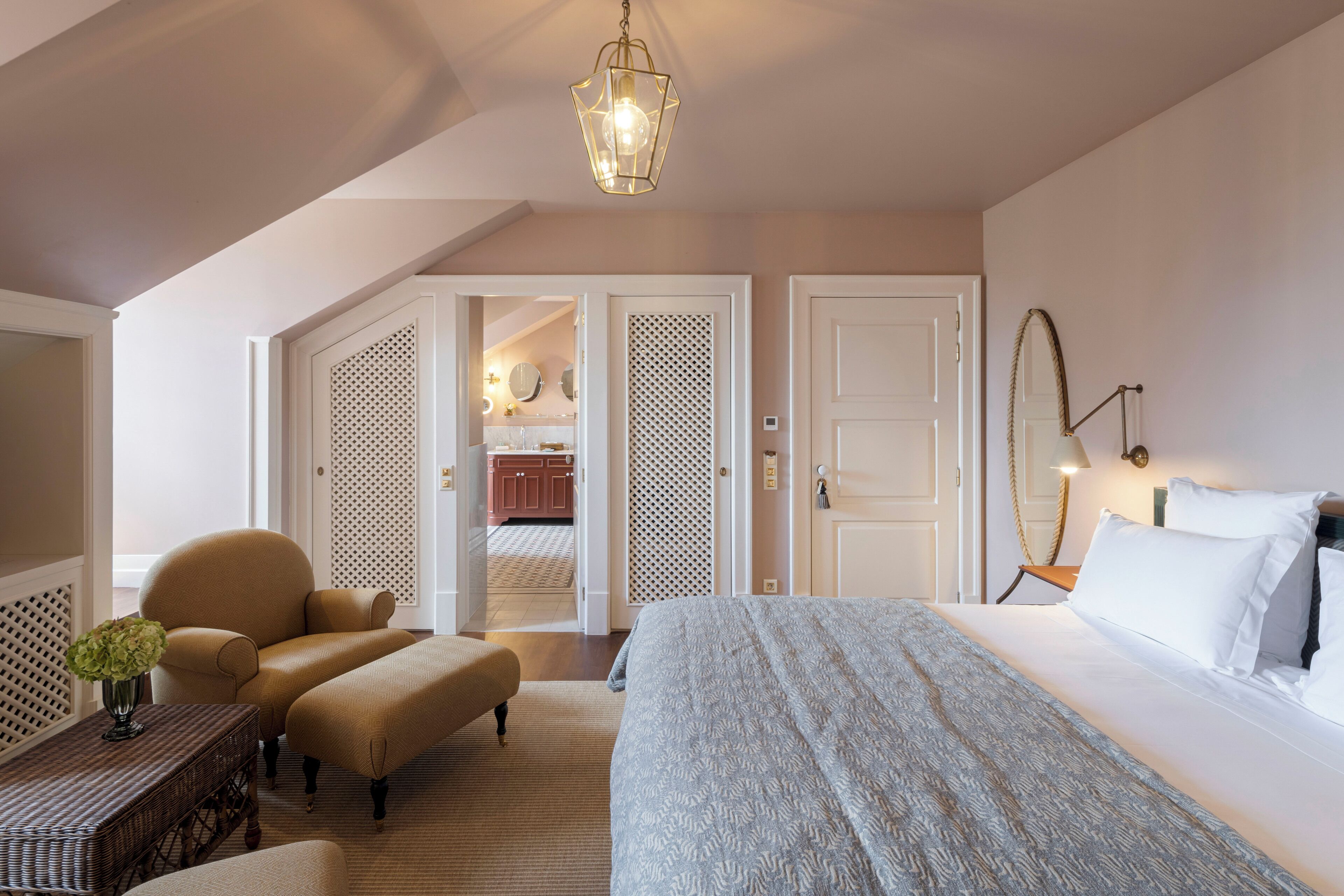 A bedroom with a large bed, a chair, a small table with a vase, and a hanging light fixture.