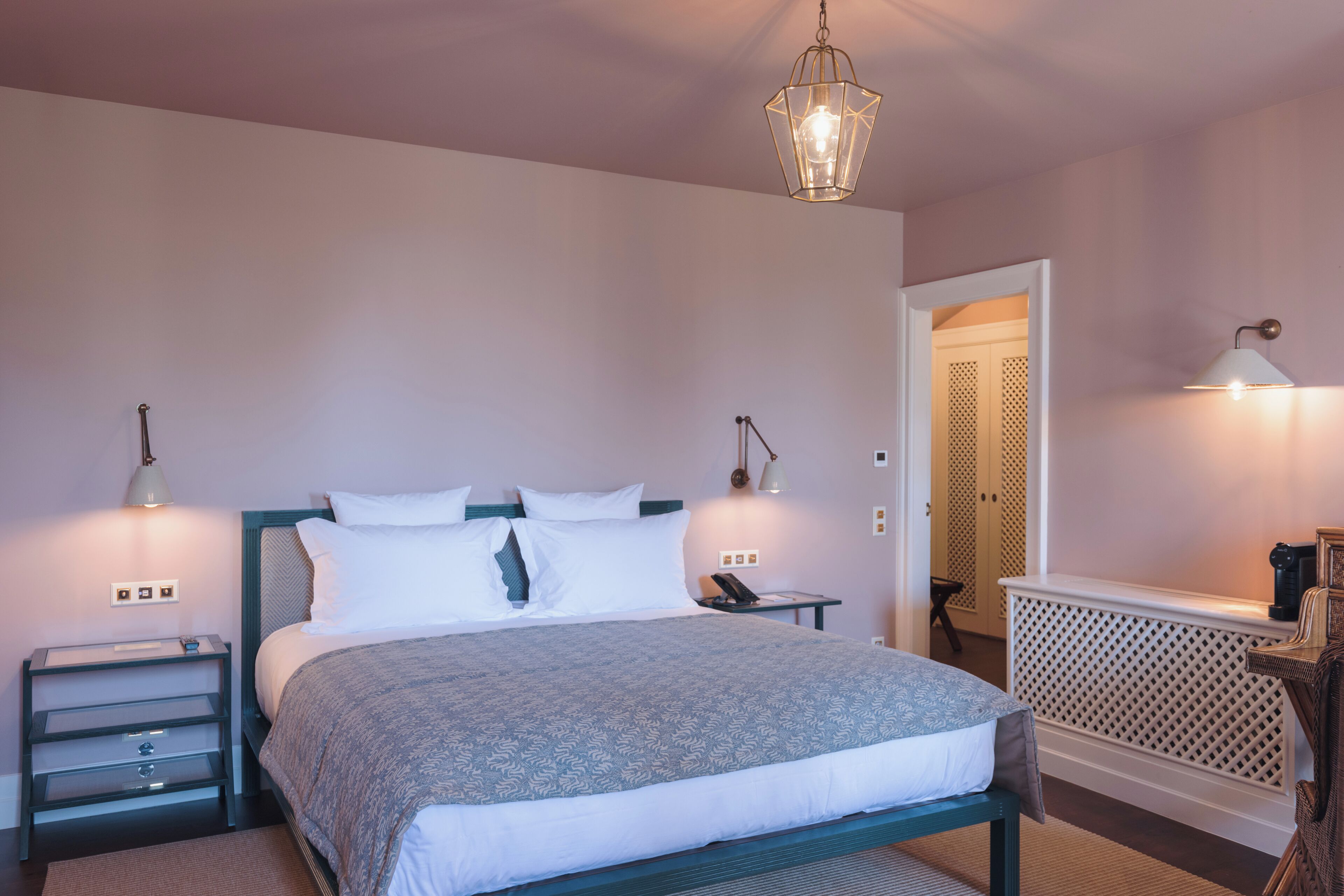 Un dormitorio ordenado con una cama, mesitas de noche, una lámpara y una puerta que da a otra habitación.