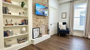 A living room with a brick wall, wooden flooring, a bookshelf, a TV mounted on the wall, a chair, and a window with curtains. - Bourbon Trio: Walkable to Restaurants, bars, shops and the Capitol building (Frankfort)