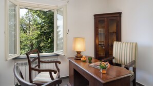 A room with a wooden chair, a table with a lamp, a cabinet, and a window with a view of trees. - Casa Sul Serra (Calci)