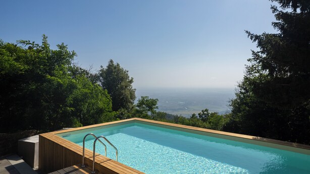 A rectangular pool with a wooden deck and metal handrails, surrounded by lush greenery and overlooking a distant landscape. - Casa Sul Serra (Calci)