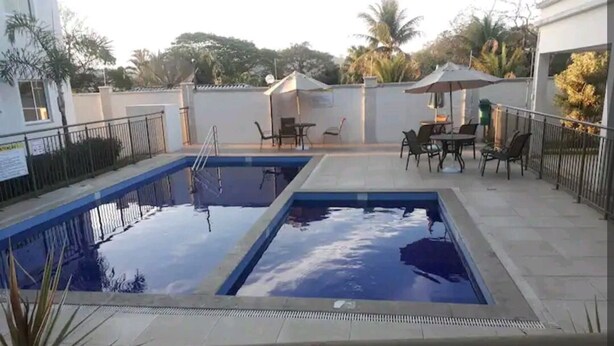 En swimmingpool med to afdelinger, omgivet af en flisbelagt terrasse med udendørs møbler og parasoller.