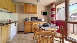 Une cuisine avec des meubles de rangement en bois, une table à manger avec des chaises, un téléviseur fixé au mur et une fenêtre avec des rideaux.