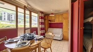 Une pièce compacte comprenant une table à manger pour deux personnes, un canapé, un lit superposé et une fenêtre donnant sur un immeuble et de la verdure.