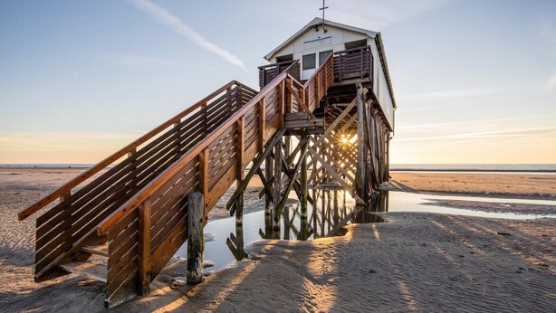 A wooden pier leading to a small building with a balcony, situated on a sandy beach with calm water in the background. - Ferienwohnung direkt am Deich in St.Peter-Ording (St. Peter-Ording)