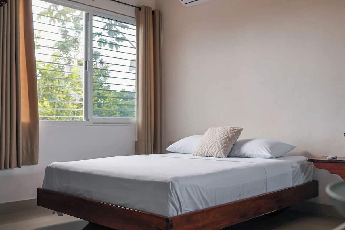 A modern bedroom with a large bed, a window with curtains, an air conditioning unit, and a wooden nightstand.