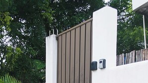 A gated entrance with a concrete wall and a metal gate, surrounded by lush greenery and palm trees. - Apartamento 22 (Bacalar)