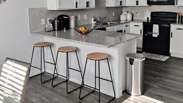 Una cocina moderna con armarios blancos, encimera de granito, microondas negro y refrigerador de acero inoxidable. Hay tres taburetes de bar y un cubo de basura.