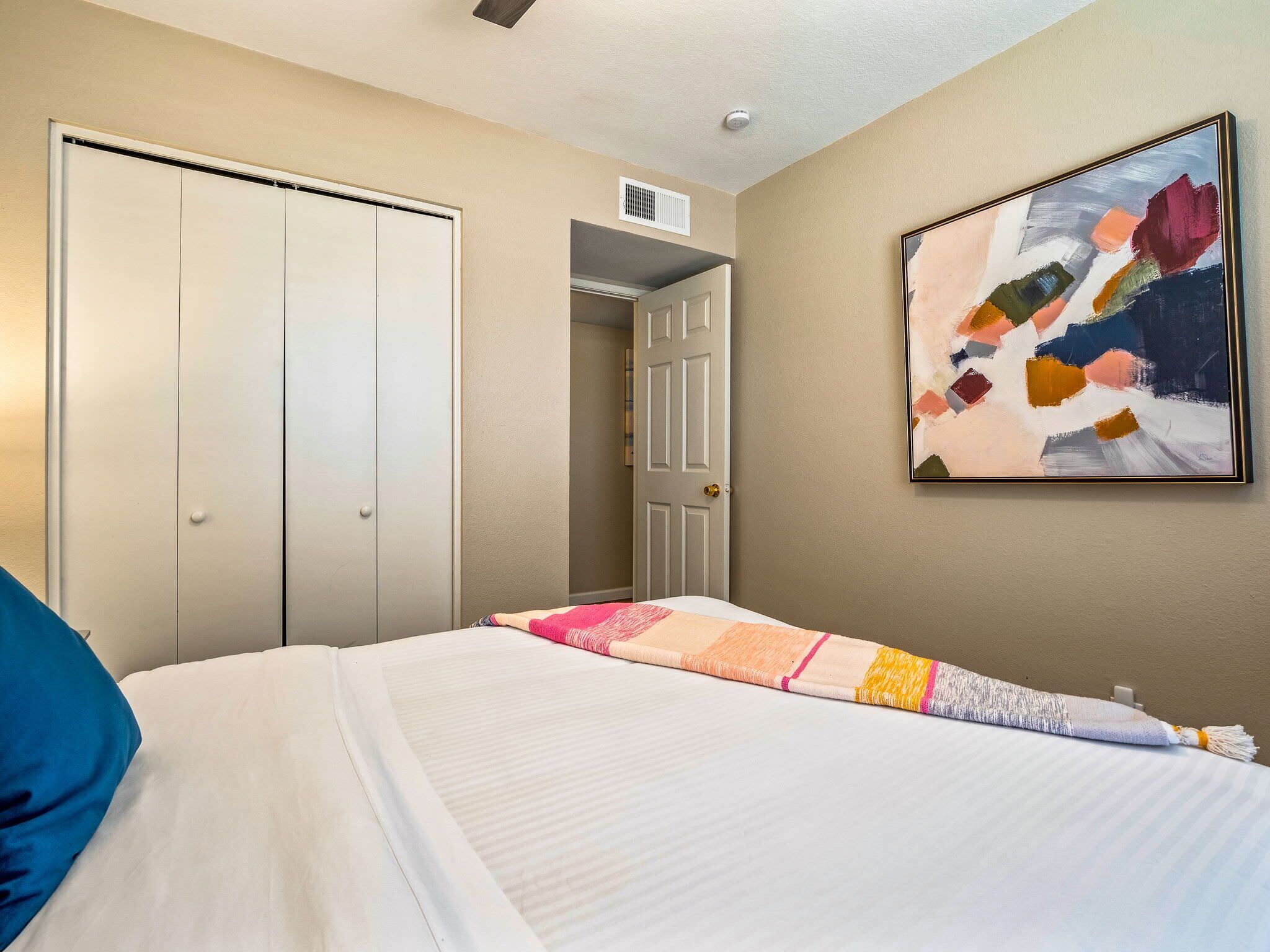 A bedroom with a bed, a wardrobe, a door leading to another room, and a painting on the wall.