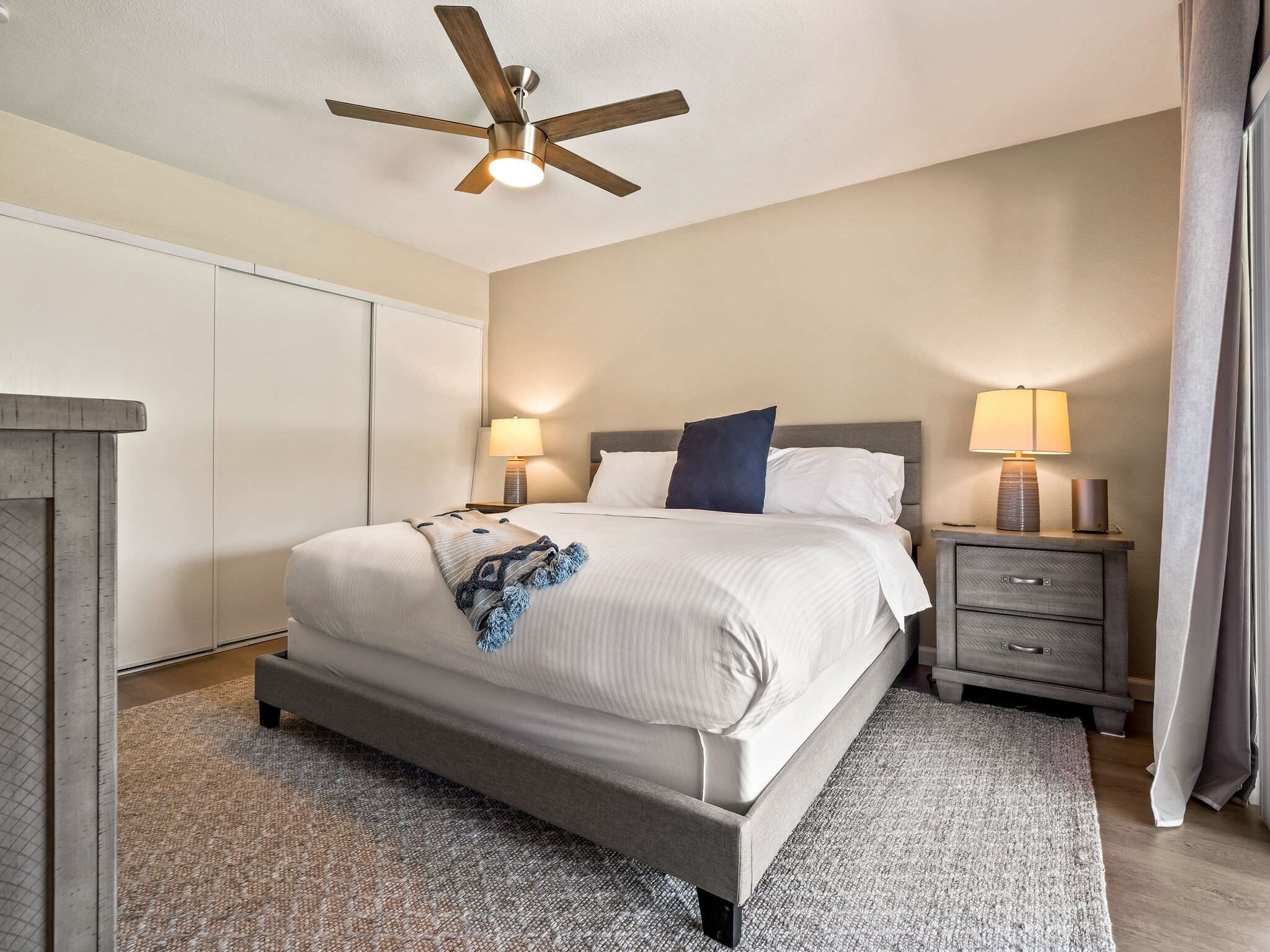A modern hotel room with a large bed, two bedside tables with lamps, a ceiling fan, and a wardrobe.